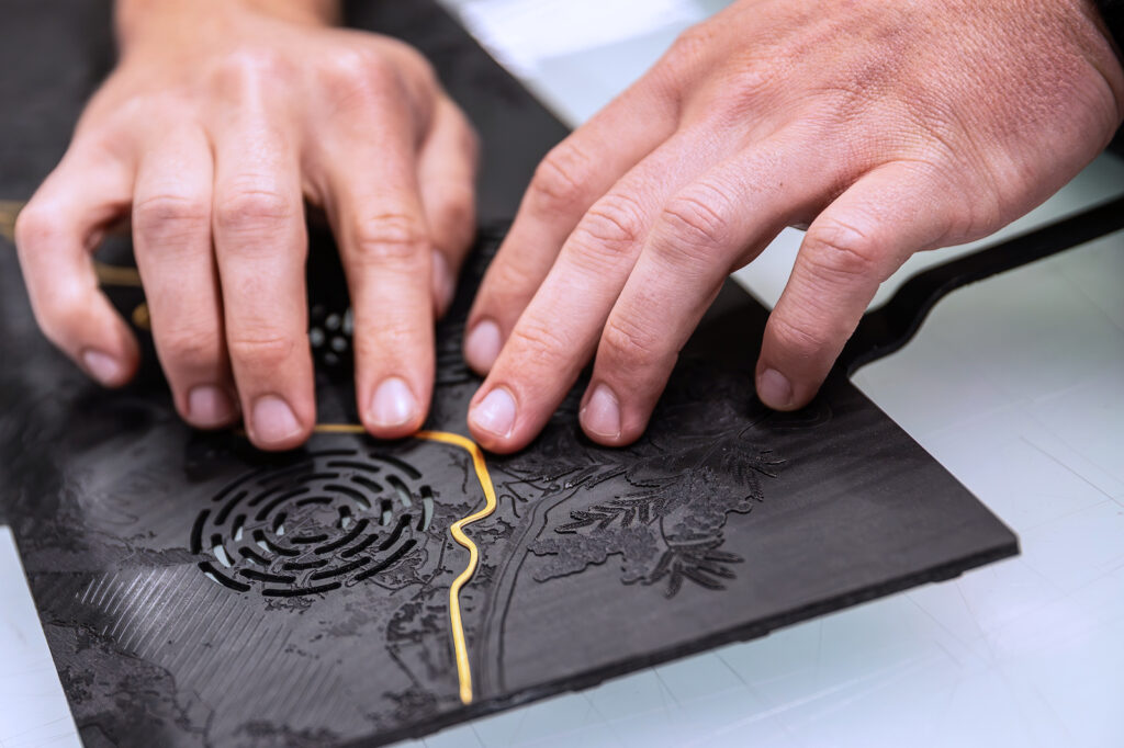 Two hands pressing gold inlay into detailed carved wood dashboard panel