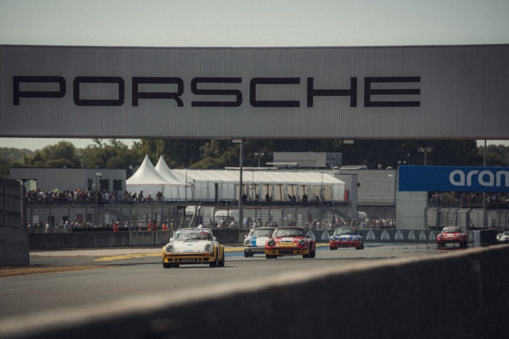 Classic Porsche race cars driving on circuit beneath Porsche bridge at Peter Auto Le Mans Classic