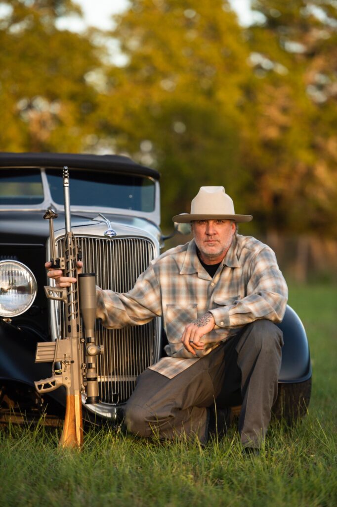 Jesse James posing with rifle in front of vintage hot rod pickup