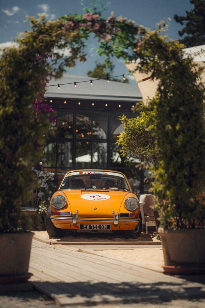 Orange classic Porsche 911 parked beneath a floral archway at a motorsport event