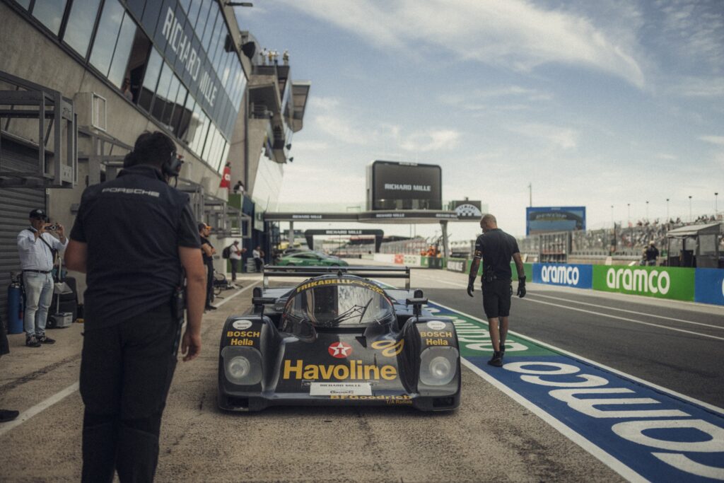 A black Havoline-liveried race car sits in the pit lane at Le Mans Classic, surrounded by team members and pit wall signage.