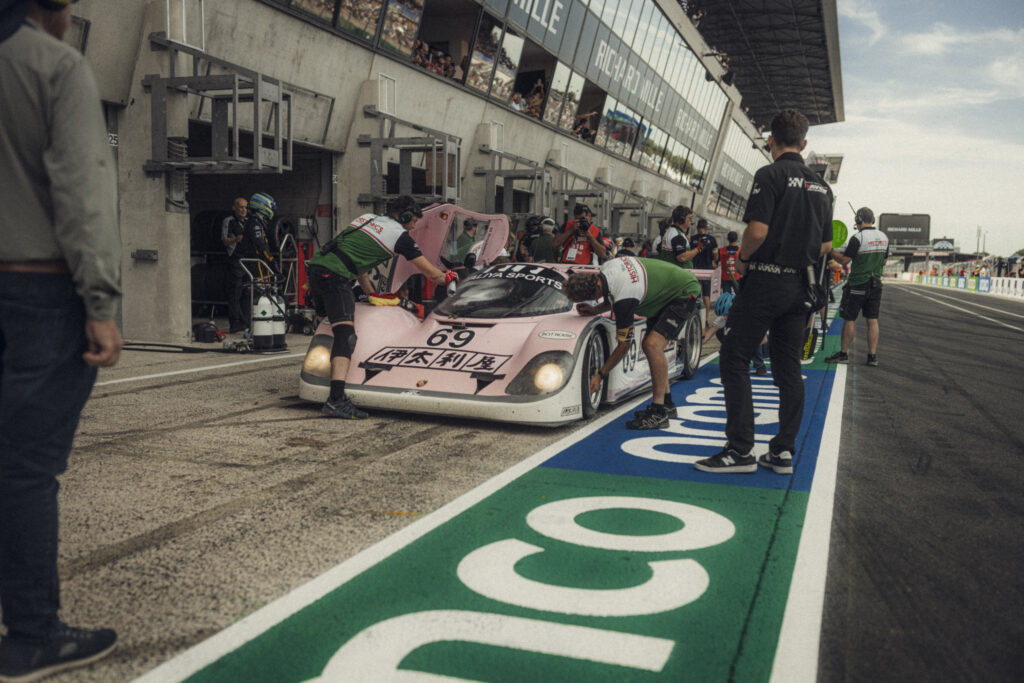 Pink historic prototype race car being worked on by crew members in the pit lane at Le Mans Classic, organised by Peter Auto