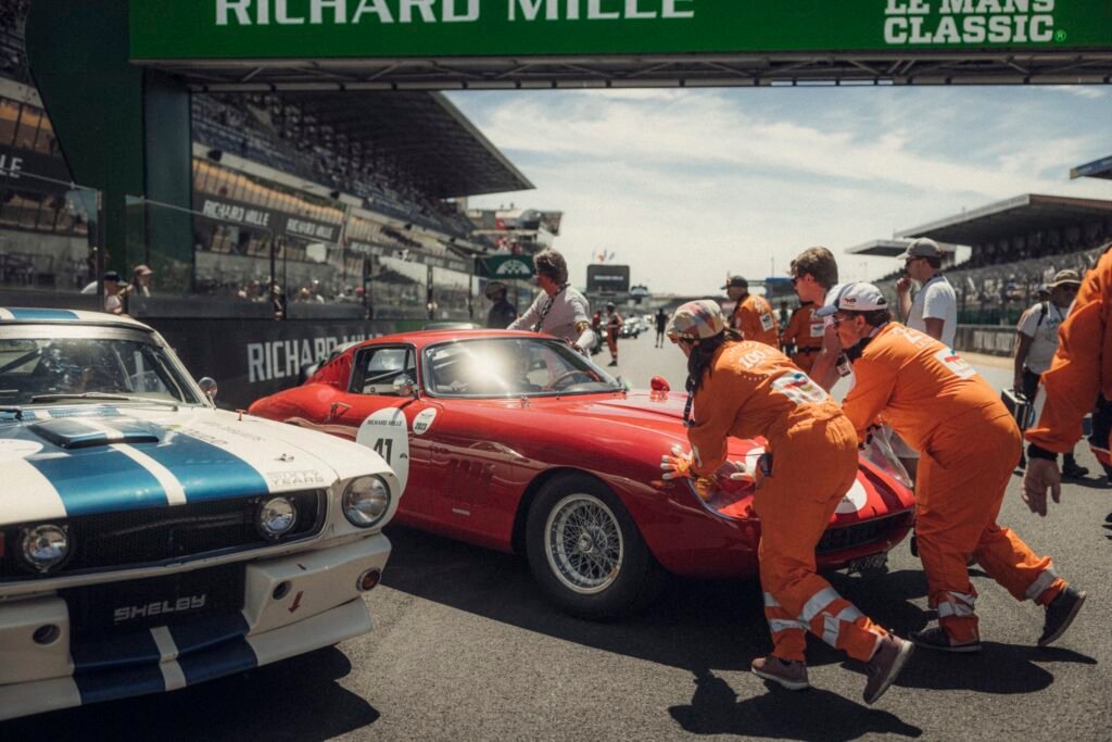 Vintage red coupe race car being pushed by marshals on the grid at Le Mans Classic, organised by Peter Auto