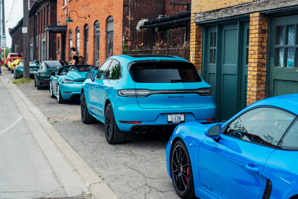 Turquoise Porsche Macan GTS parked in a lineup of Porsche sports cars on a brick-lined street at a Kaleidoscope car gathering