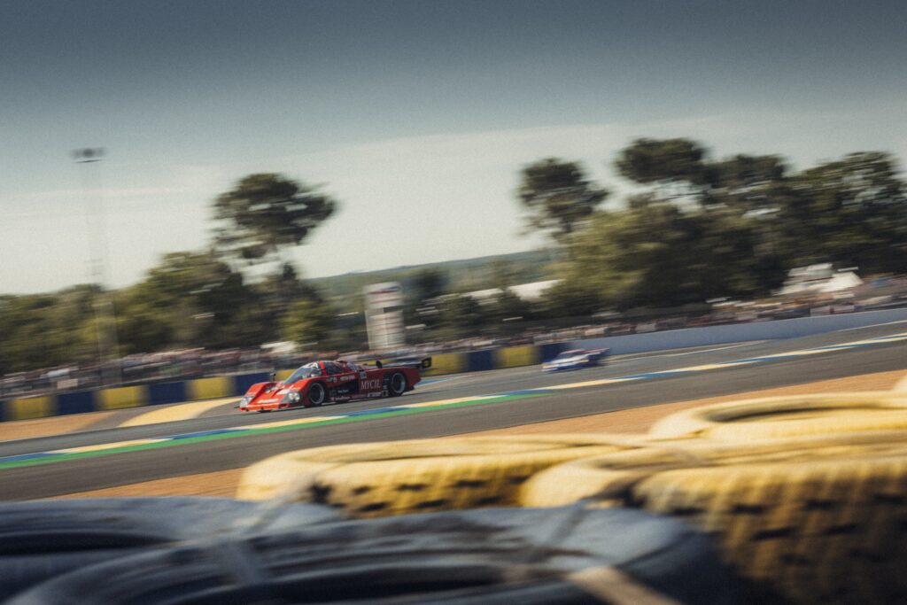 Red prototype race car on track seen behind tire barriers at Classic Le Mans