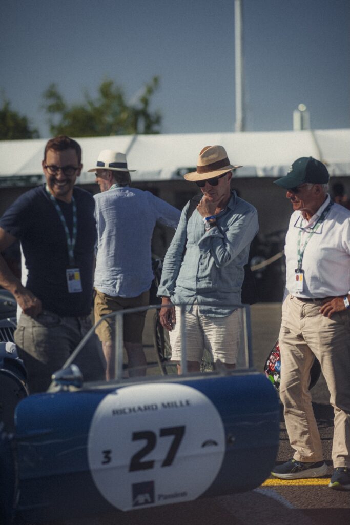 Group of spectators standing beside a blue vintage race car in the paddock
