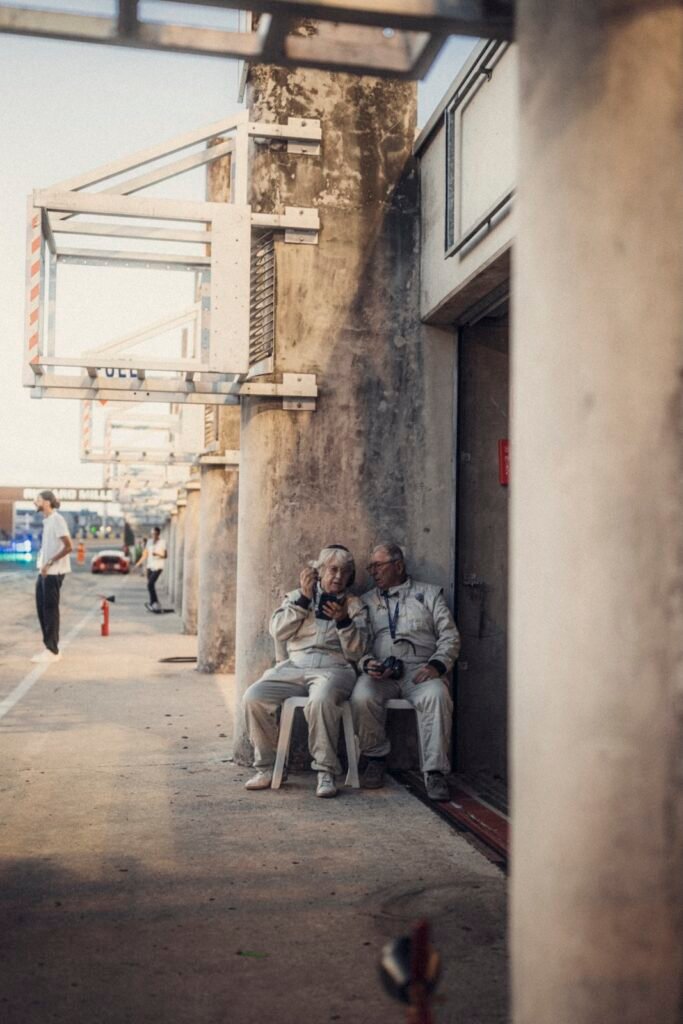 Two vintage race drivers in white suits sitting on chair beside pit garage wall at Le Mans Classic