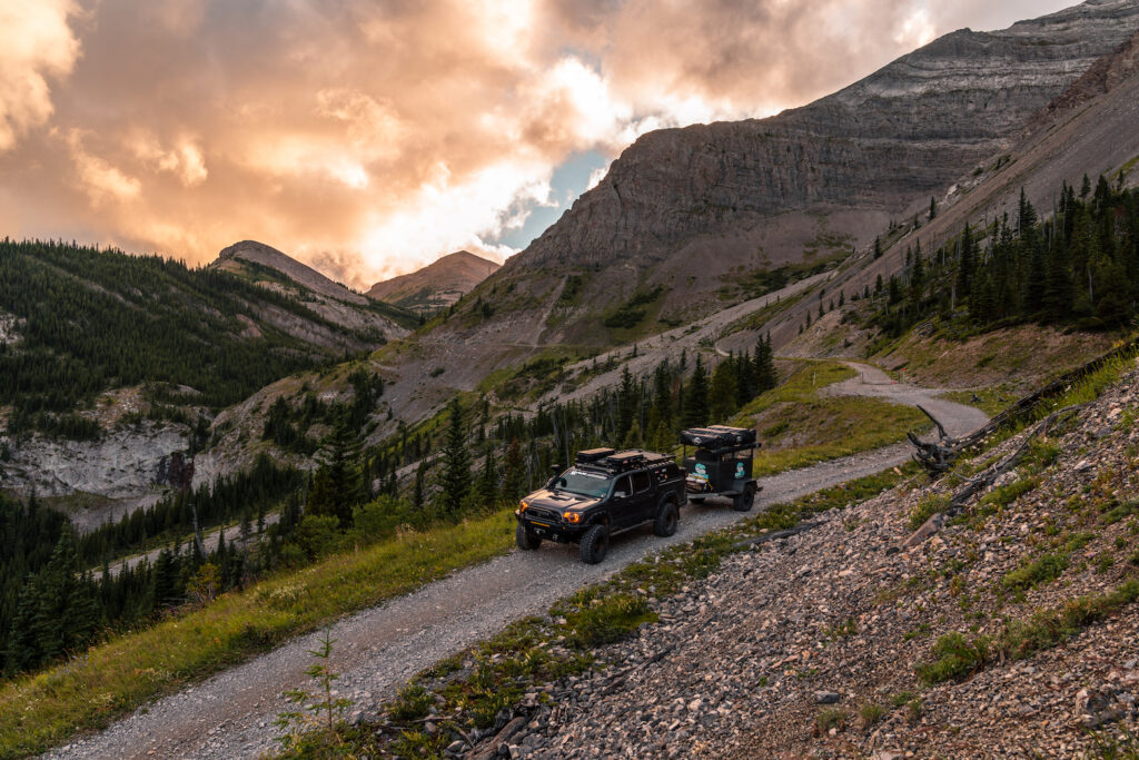Toyota Tacoma towing an overland trailer on a high alpine mountain road at sunset