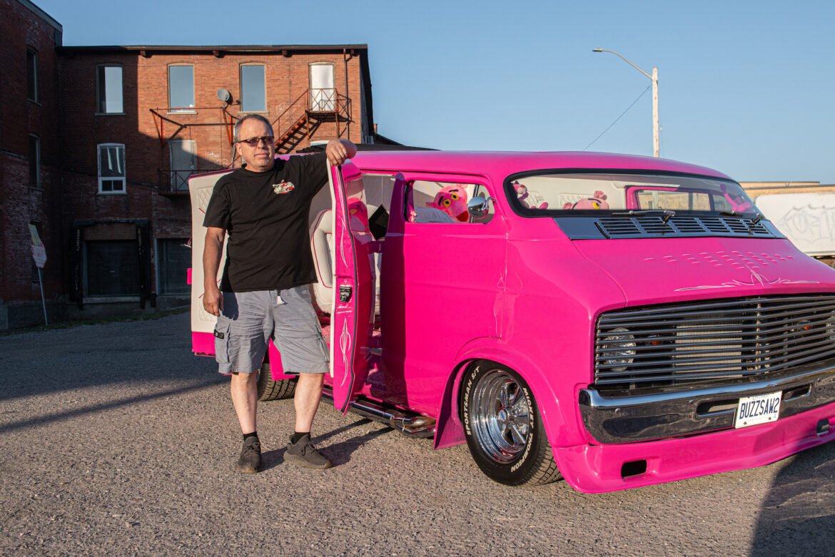 Ken McCarl and Buzz Saw 2.0 Ken McCarl poses beside Buzz Saw 2.0, a bright pink custom Dodge van with a chopped roof, chrome grille and Pink Panther-themed interior, parked outside a brick industrial building at sunset.
