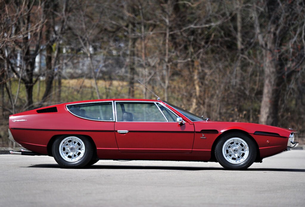 Side profile of a red 1971 Lamborghini Espada parked on a road, showing the classic four-seat grand tourer's long, low silhouette against a backdrop of leafless trees.