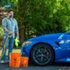 Thomas Holland washes a blue Alfa Romeo Giulia with a garden hose beside two orange buckets.